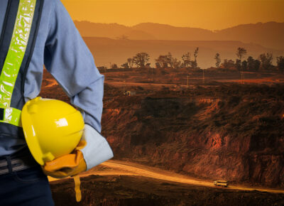 Engineer holding a yellow helmet for the safety of workers on a background of coal mining trucks are driving on the road. The sunset